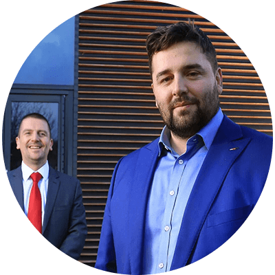 A man with brown hair and a brown bear smiling with a blue suit on and a man with a black suit stood.