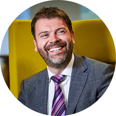 A man with short brown hair and a beard smiling wearing a grey suit with a white shirt and colourful purple tie.