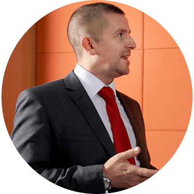 A man with short brown hair wearing a black suit in a discussion.