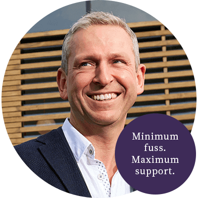 A man with short grey hair smiling with a black suit on with a purple circle in front saying minimum.