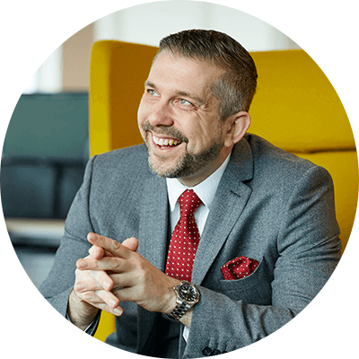 A man with short grey hair wearing a grey suit while smiling sat on a yellow chair.