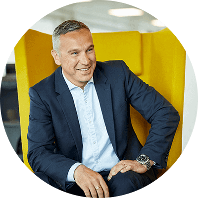 A man with short grey hair with a navy blue suit on smiling on a yellow seat.