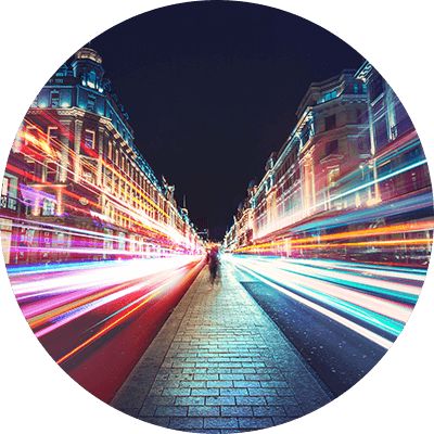 A path in the middle of a road with buildings on either side and blurred lights of cars going past.