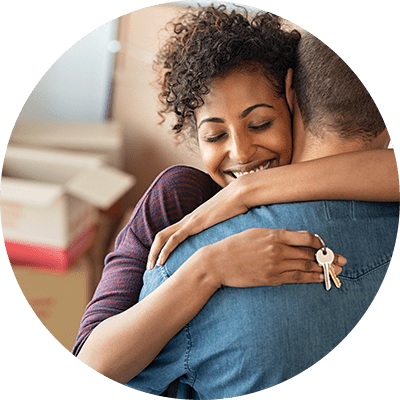 A woman hugging her husband with a set of keys in her hand and boxes behind them.