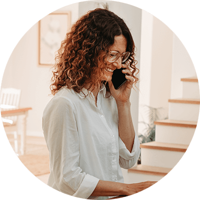 A woman with brown curly hair with a white blouse on smiling on the phone.