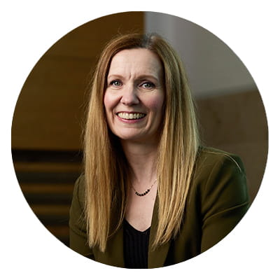 A woman with long blonde hair smiling wearing a brown blazer and black blouse.