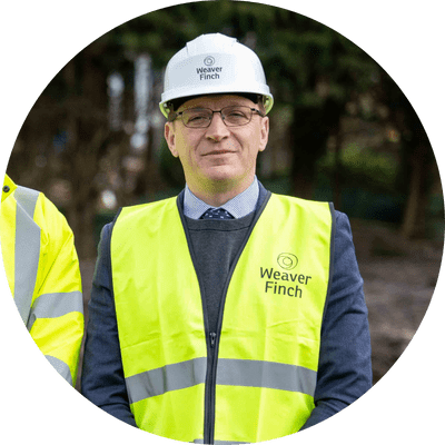 Andrew a man with glasses on building site wearing smart shirt and tie under a hi vis vest and a white hard hat.
