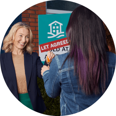 A smart female landlord handing keys to a new tenant in a denim jacket with black hair with purple streaks outside of a red brick house with a Let Agreed sign in front of it.