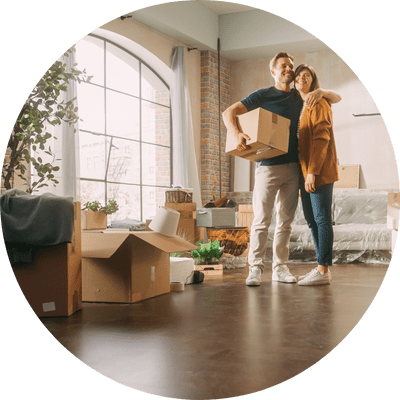 A happy young couple moving into the brightly window-lit front room of their new home. The smiling man is holding a box of belongings with his other arm around the woman.
