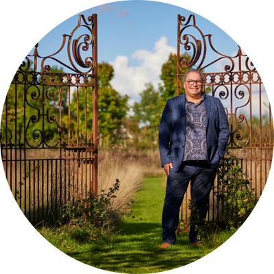 Ken a man in patterned shirt under a casual blue suit stood next to ornate iron gates.