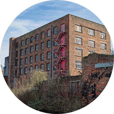 Historic brick mill building with red fire escape near water on a clear day. Similar properties have been converted into new commercial premises, including offices and mixed-use spaces.