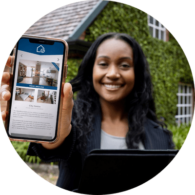 Professional woman showing a property app on her phone with a house covered in green vines behind her.