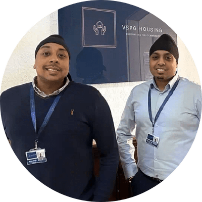 Two men wearing blue smiling in front of a sign.