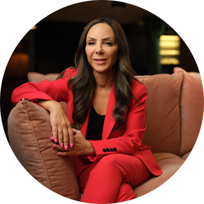 A mid-shot of Tanya Elmaz with long brown hair smiling whilst sat on a couch in a bright coloured suit.