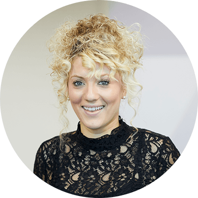 A woman with curly blonde hair in a ponytail smiling wearing a black lacey dress.