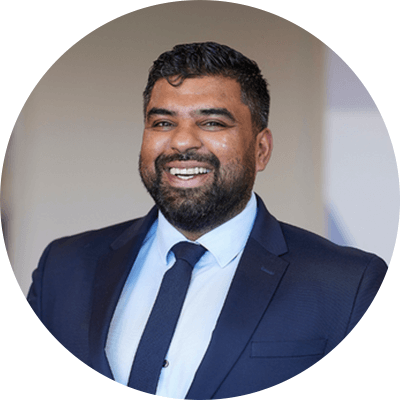 A man with short black hair and a black beard smiling wearing a navy blue suit.