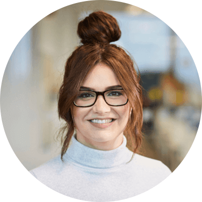 A woman with ginger hair in a bun smiling with glasses on and a white turtleneck.