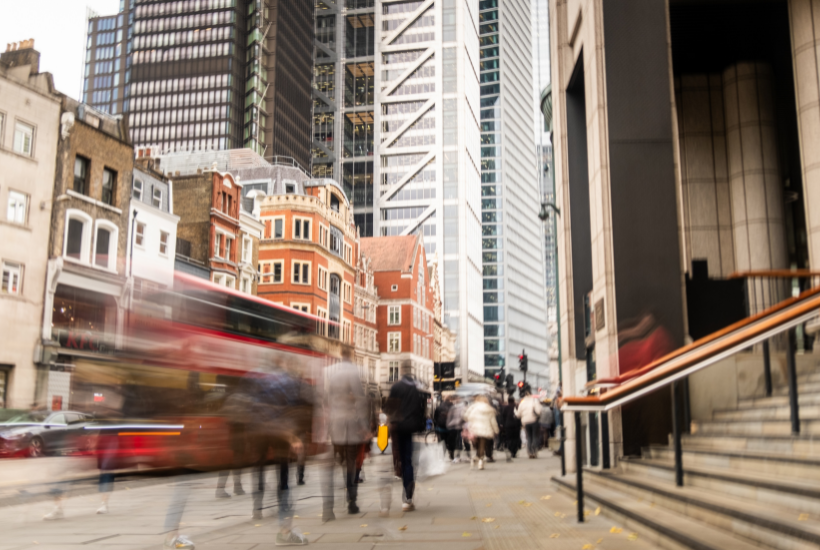 Busy UK street with shoppers and a bus partially blurred to show movement, with large office buildings in the background.