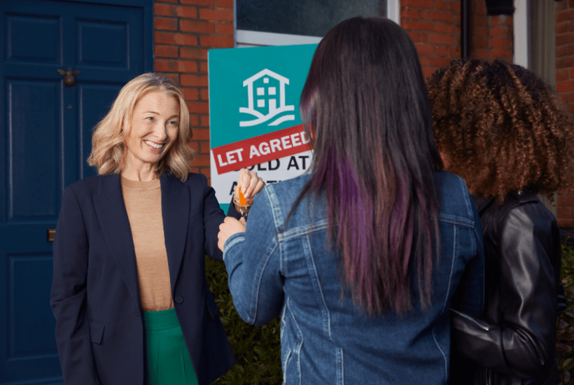 Older female landlord with blonde hair smiling and handing door keys to a young female couple outside of a house with a blue door with a Let Agreed sign in the garden.