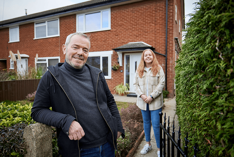 man with grey hair stood in front of a garden gate next to a woman in front of a house - l