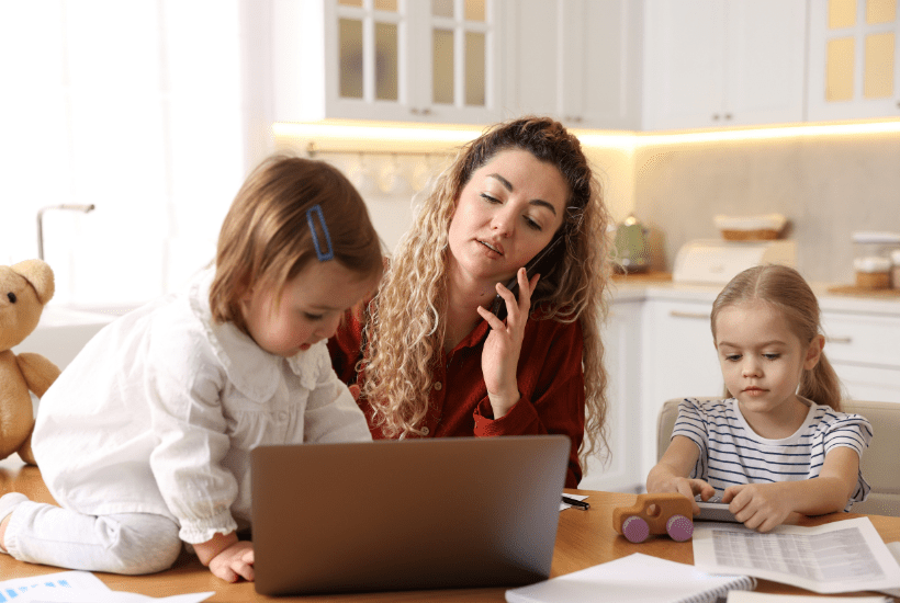 A busy and frustrated mum trying to work on mobile phone in a kitchen, with a young child colouring next to her and a toddler playing with her mum's laptop.