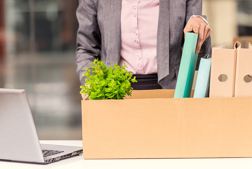 woman in grey jacket putting a file in a cardboard box containing files and a plant - l