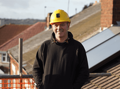 A man wearing all black with a yellow hardhat on on top of a roof.