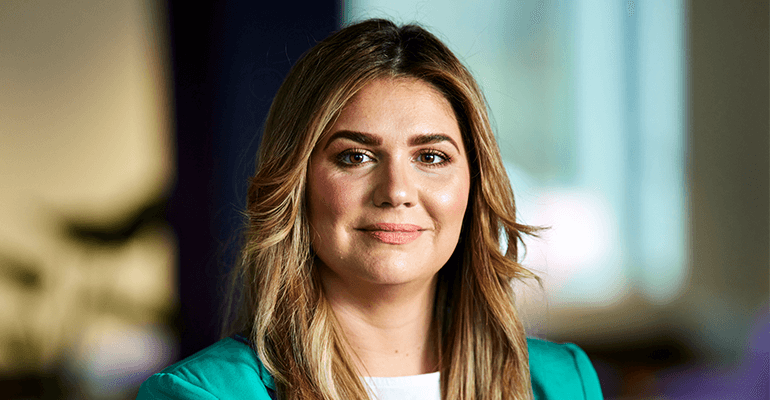 A close up of a woman with long light brown hair smiling wearing a green blazer and white top.