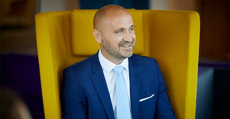 A middle-aged bald man with a beard smiling sat on a yellow seat.