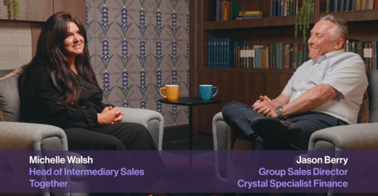 A man and woman sat on comfortable chairs talking in front of a bookcase and fireplace.
