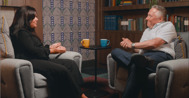 A man and woman sat on comfortable chairs talking in front of a bookcase and fireplace.