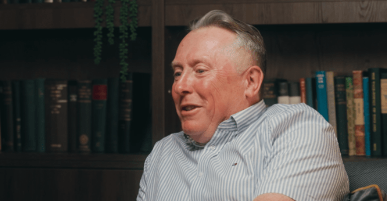 A middle-aged man in a white short sleeve shirt sat in front of a bookcase.