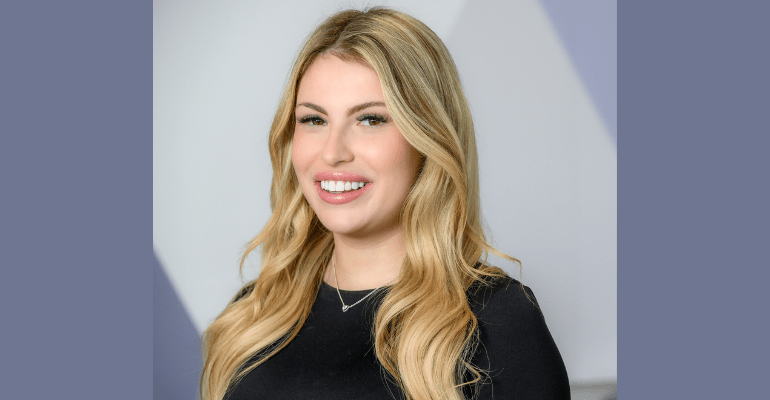 Young blonde professional woman smiling in front of a neutral office background, wearing a black top with a visible necklace.