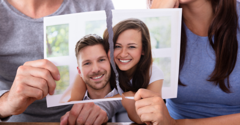 Close up of a ripped picture showing a previously happy young couple smiling. The couple are holding separate section of the picture and look unhappy, signalling separation or divorce.