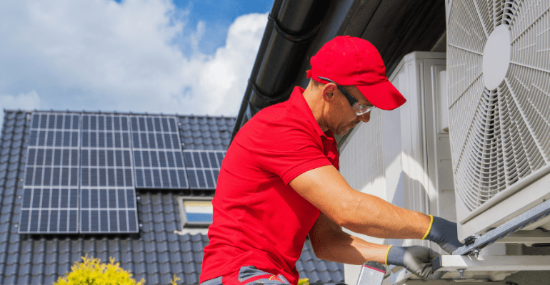 Engineer in red overall installs a heat pump on the outside of a house.