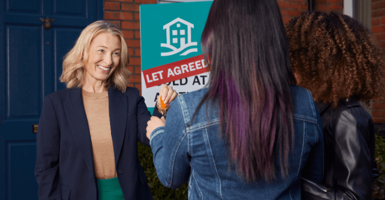 Older female landlord with blonde hair smiling and handing door keys to a young female couple outside of a house with a blue door with a Let Agreed sign in the garden.