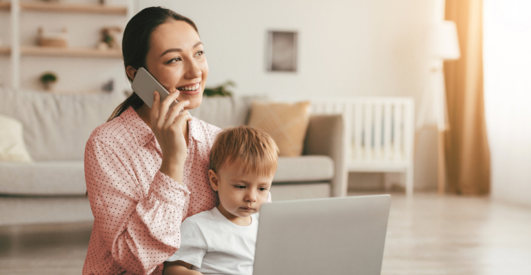 A happy mum on a phone sat on the floor in a living room with her child and laptop on her lap.