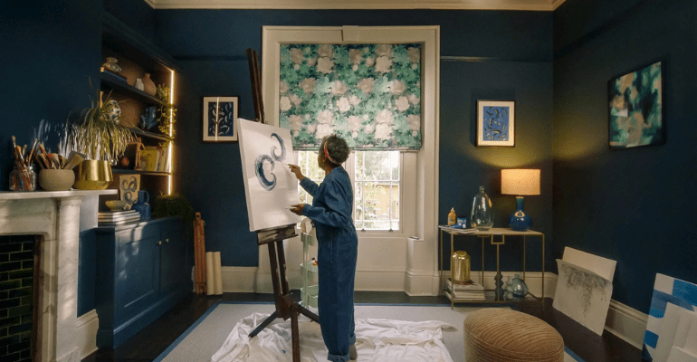 Older black lady painting in a stylish front room in front of a window with floral patterned blind. Credit - Hillarys Blinds.