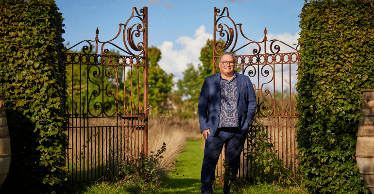 Ken a man in patterned shirt under a casual blue suit stood next to ornate iron gates.