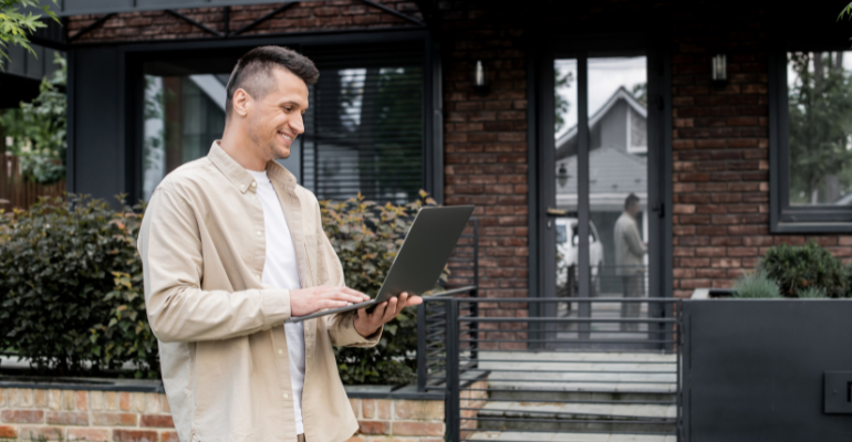 A happy landlord dressed casually reviewing a laptop outside of a rental property.