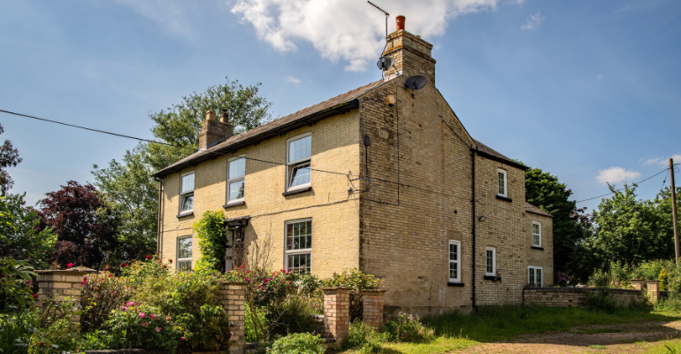 A large yellow brick farmhouse in the countryside with a beautiful but slightly overgrown garden under blue skies.