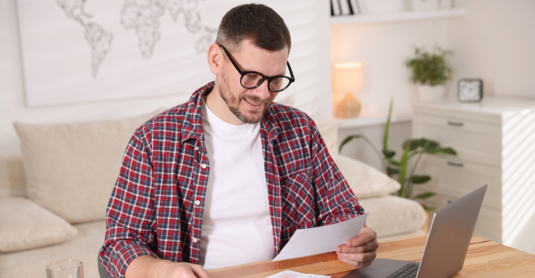 A smiling man in glasses wearing a red, black and white check shirt sat in a living room reviewing finances.