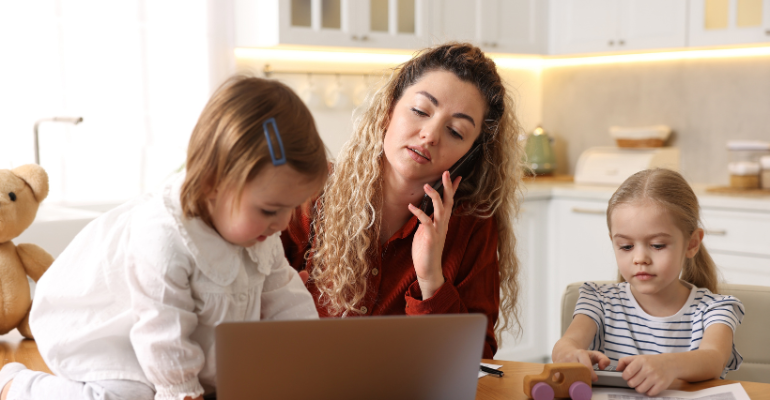 A busy and frustrated mum trying to work on mobile phone in a kitchen, with a young child colouring next to her and a toddler playing with her mum's laptop.