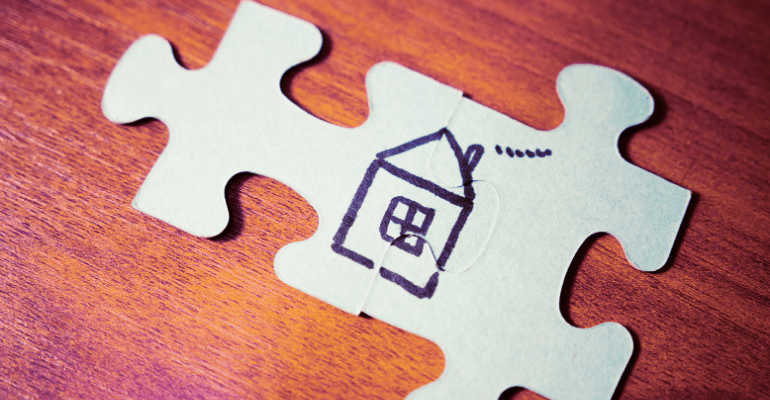 A puzzle piece with a basic hand drawn picture of a house on it, on a wooden varnished surface.