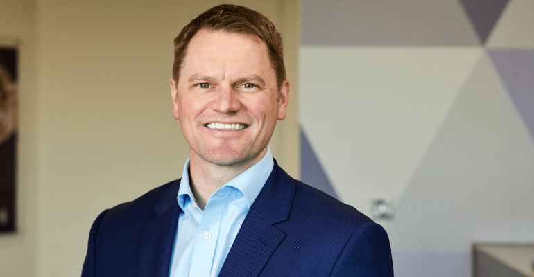 Middle aged man with dark blonde hair in a dark blue suit and light blue shirt smiling in an office setting.