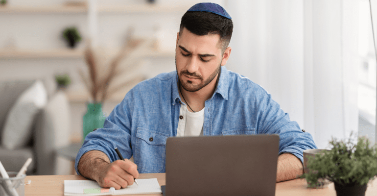 Young Jewish male business owner working at a desk, writing on a pad in front of a laptop.