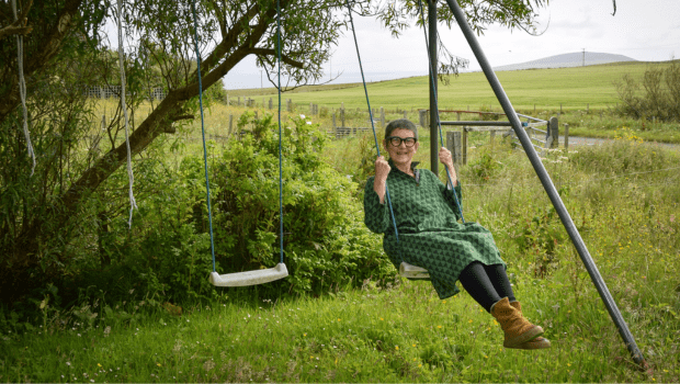 Older woman in green dress on a swing with countryside view.