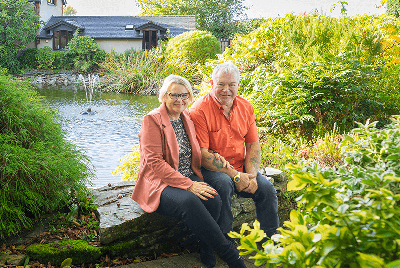 An older couple sat on a stone wall in front of a pond next to their house.