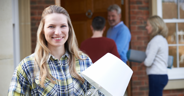 Young blonde woman smiling and holding a lamp, stood outside a houses she's moving in to, with older relatives helping in the background.