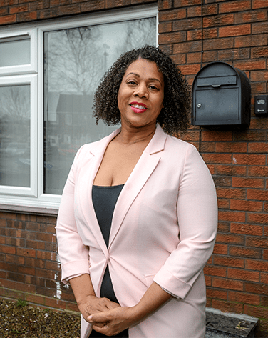 A woman standing outside of a house smiling whilst wearing a pink blazer and black top.